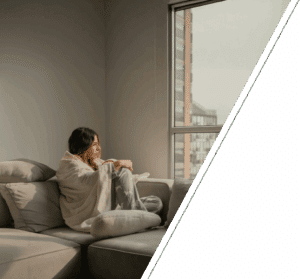 A high-functioning adult experiencing depression and fatigue from chronic illness sits alone in a modern apartment, gazing out at the gray sky and city buildings. This emotional image captures the feeling of being trapped in one’s body, navigating the hidden struggles of anxiety from chronic pain, grief, and identity crisis linked to chronic illness. The setting conveys the invisible nature of illness, where someone may look fine but feel awful inside, emphasizing mental health support for chronic illness, therapy for chronic pain, and compassionate care for those powering through while feeling like a burden.