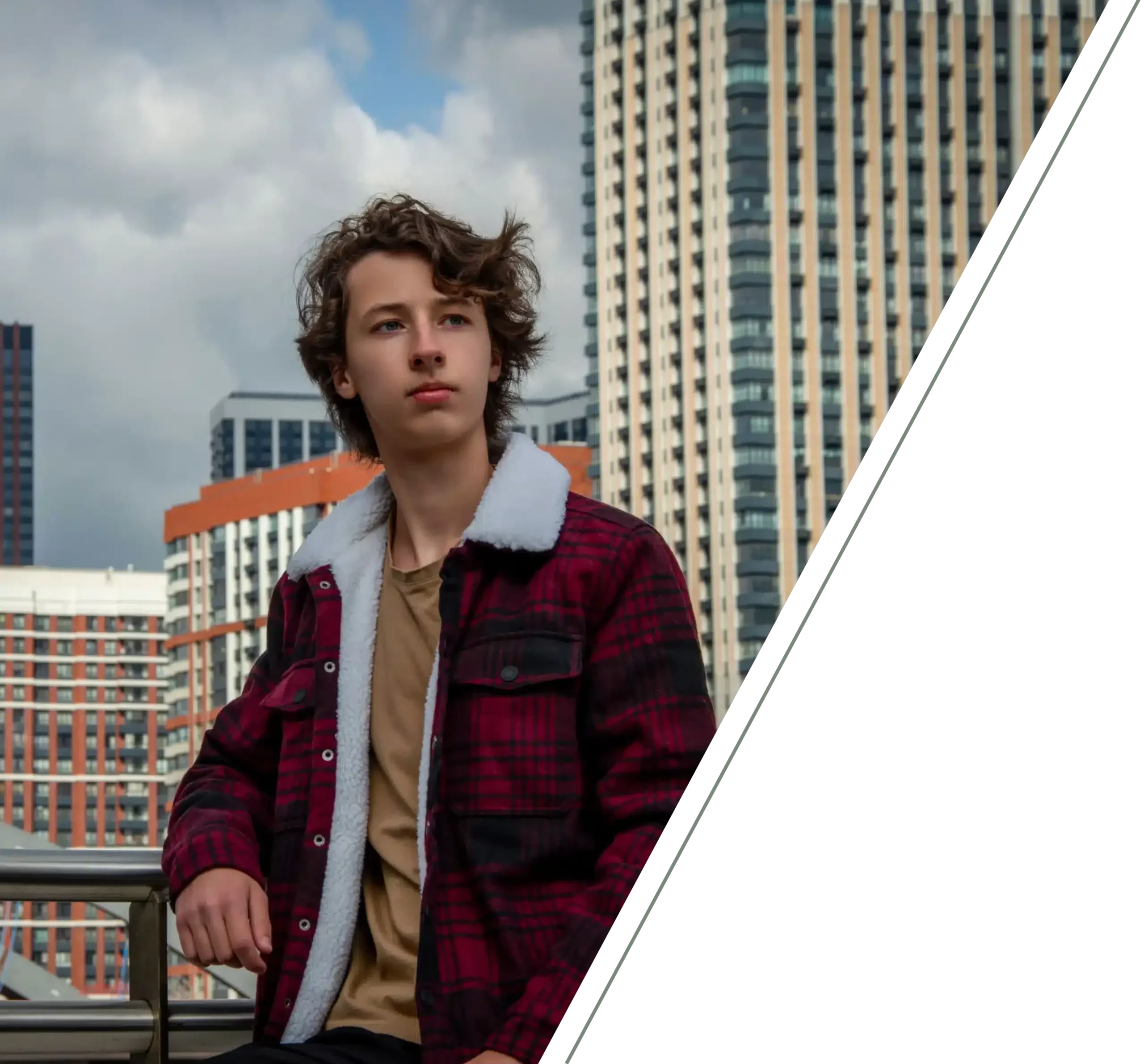 Young man with wavy hair wearing a red sherpa-lined plaid jacket posing outdoors with modern city skyscrapers in the background.